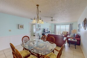 A dining area with a glass-top table surrounded by wooden chairs, a ceiling fan, and a view of the living room.