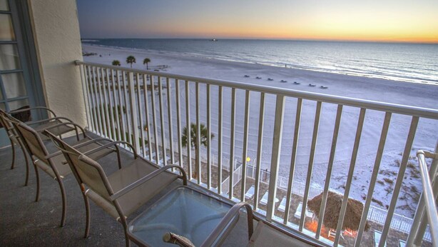 A balcony with a glass table and chairs, overlooking a beach with a fence and palm trees.