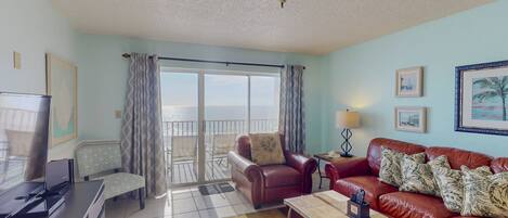 A living room with a TV, a sofa, a coffee table, and a balcony with a view of the sea.