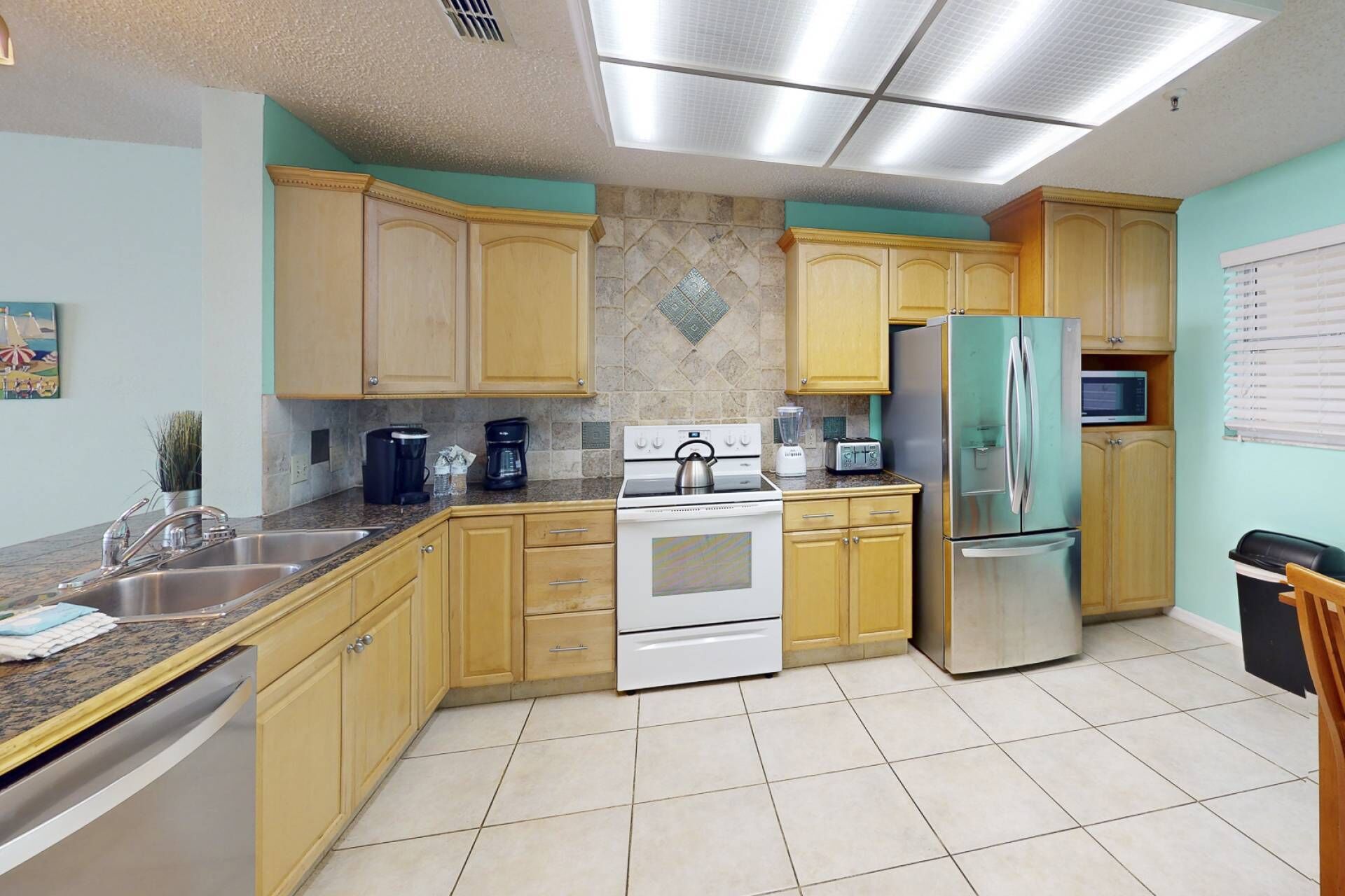 Una cocina con armarios de madera clara, una estufa blanca, un refrigerador de acero inoxidable y piso de baldosas.