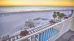 A beach view from a balcony with a pool, palm trees, and a red vehicle on the sand.