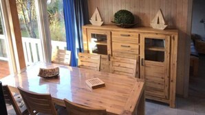Une table à manger en bois, accompagnée de chaises, un buffet avec des portes en verre, et une vue sur l’extérieur par une fenêtre.