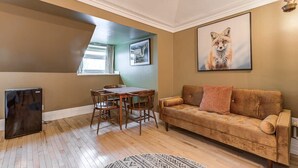 A living room with a brown sofa, a dining table with chairs, a black refrigerator, and a painting of a fox on the wall.