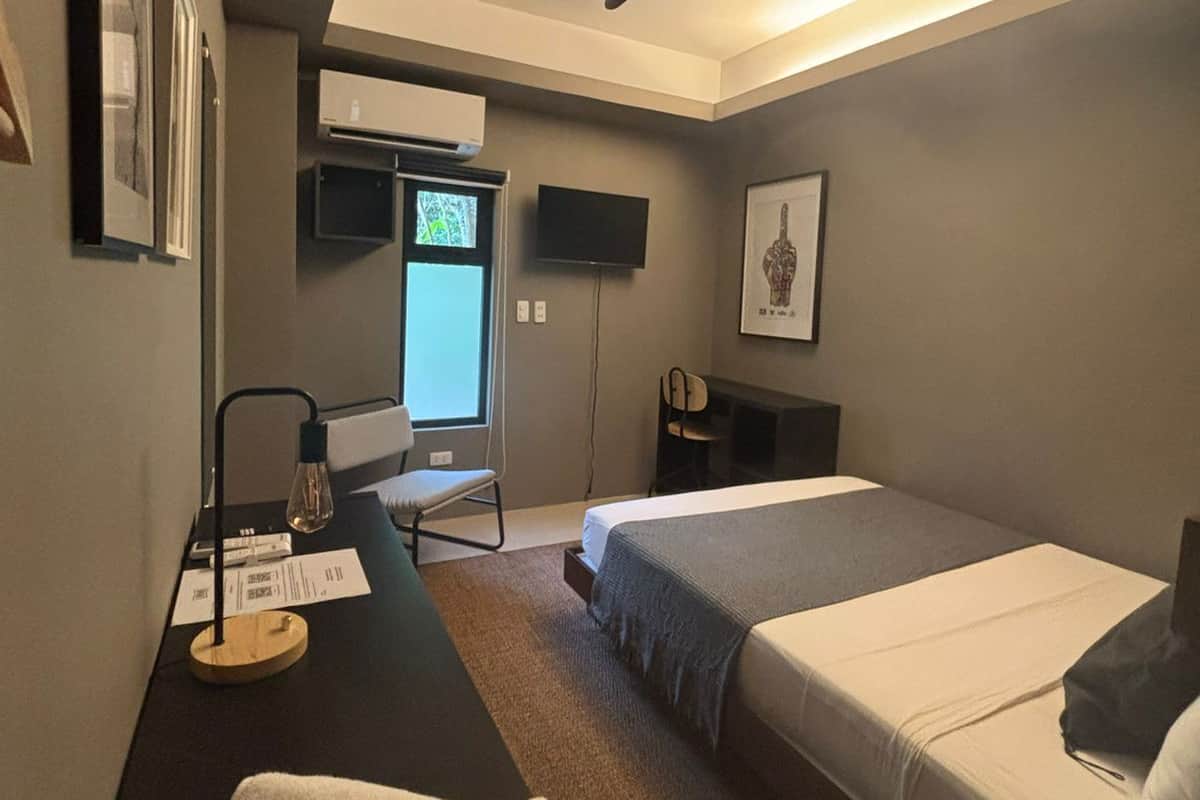 A modern hotel room with a desk, two beds, a ceiling fan, a TV, and a window.