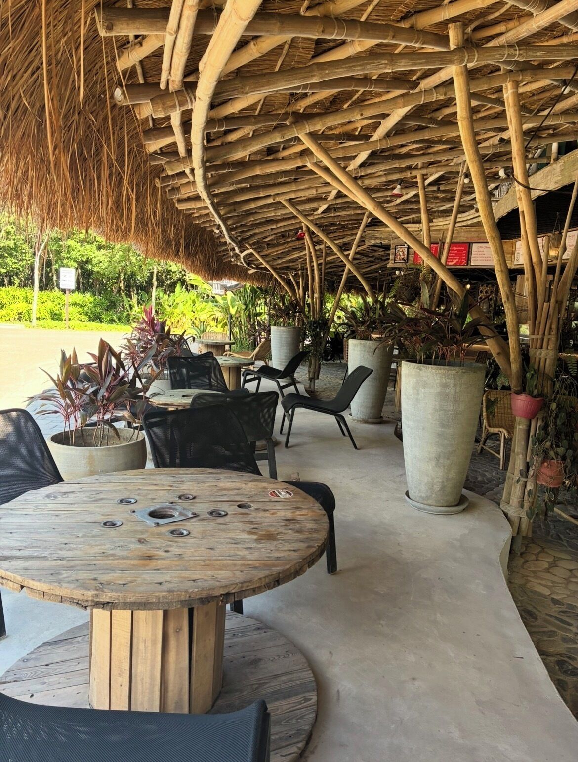Un área de estar al aire libre con mesas y sillas de madera, rodeada de plantas en macetas y una estructura de bambú en lo alto.