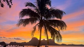 Ein Strand mit einer Palme, Menschen sitzen und gehen, und ein Sonnenuntergang im Hintergrund.