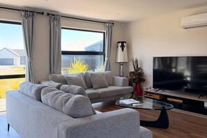 A modern living room with a grey sofa, a glass coffee table, a flat-screen TV, and a large window with curtains.