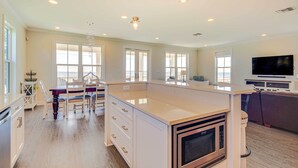 A spacious kitchen with a large island, white cabinetry, and a built-in microwave. There are multiple windows providing natural light, and a flat-screen TV mounted on the wall. - Walk to Beach: Oceanfront Home w/ Outdoor Kitchen (Waveland)