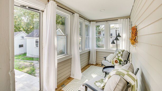Ein gemütliches Zimmer mit Blick auf einen Garten, ein Fenster mit weißen Vorhängen, ein Sessel mit Kissen und eine Vase mit Blumen.