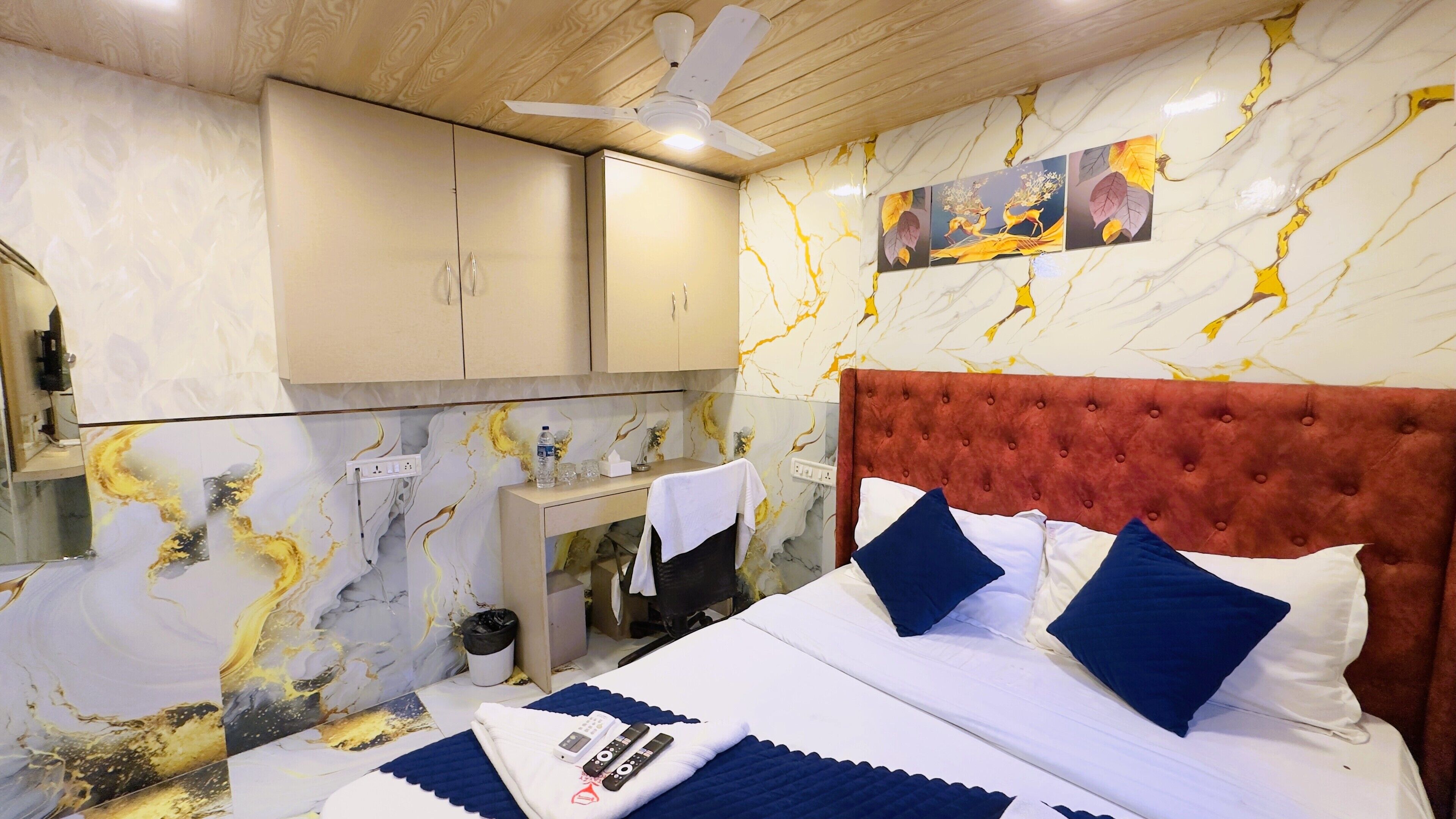 A hotel room with a marble-patterned wall, a tufted red headboard, a bed with white and blue bedding, and a small desk with a mirror and a bottle of water.