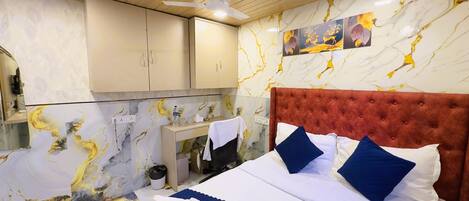 A hotel room with a marble-patterned wall, a tufted red headboard, a bed with white and blue bedding, and a small desk with a mirror and a bottle of water.