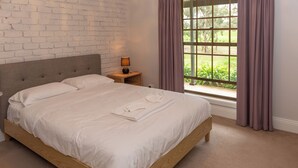 A neatly made bed with white linens and a folded towel, a wooden nightstand with a lamp, and a window with curtains. - Furche Barossa Vineyard & Fireplace  (Stone Well)