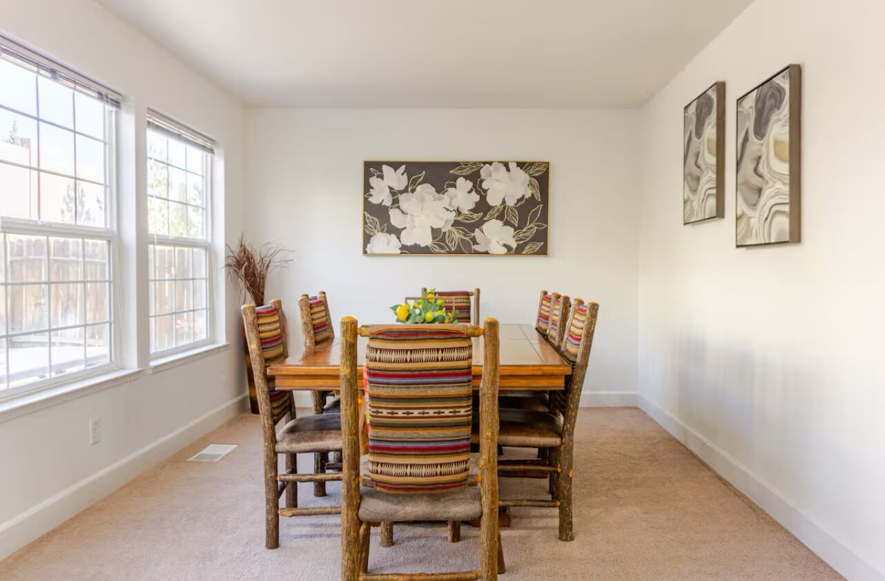 Une salle à manger avec une table et des chaises en bois, un tableau floral accroché au mur et de grandes fenêtres.