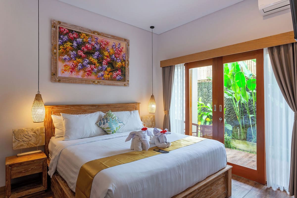 A hotel room with a large bed, wooden headboard, two bedside lamps, a painting of flowers, and a sliding glass door to a garden.
