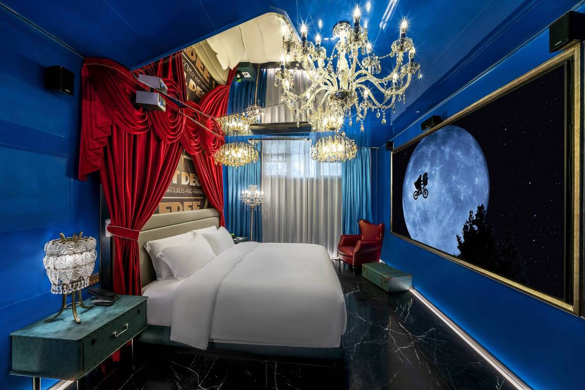 Een slaapkamer met een groot bed, rode gordijnen, een kroonluchter en een tv aan de muur met een maanlandschap.
