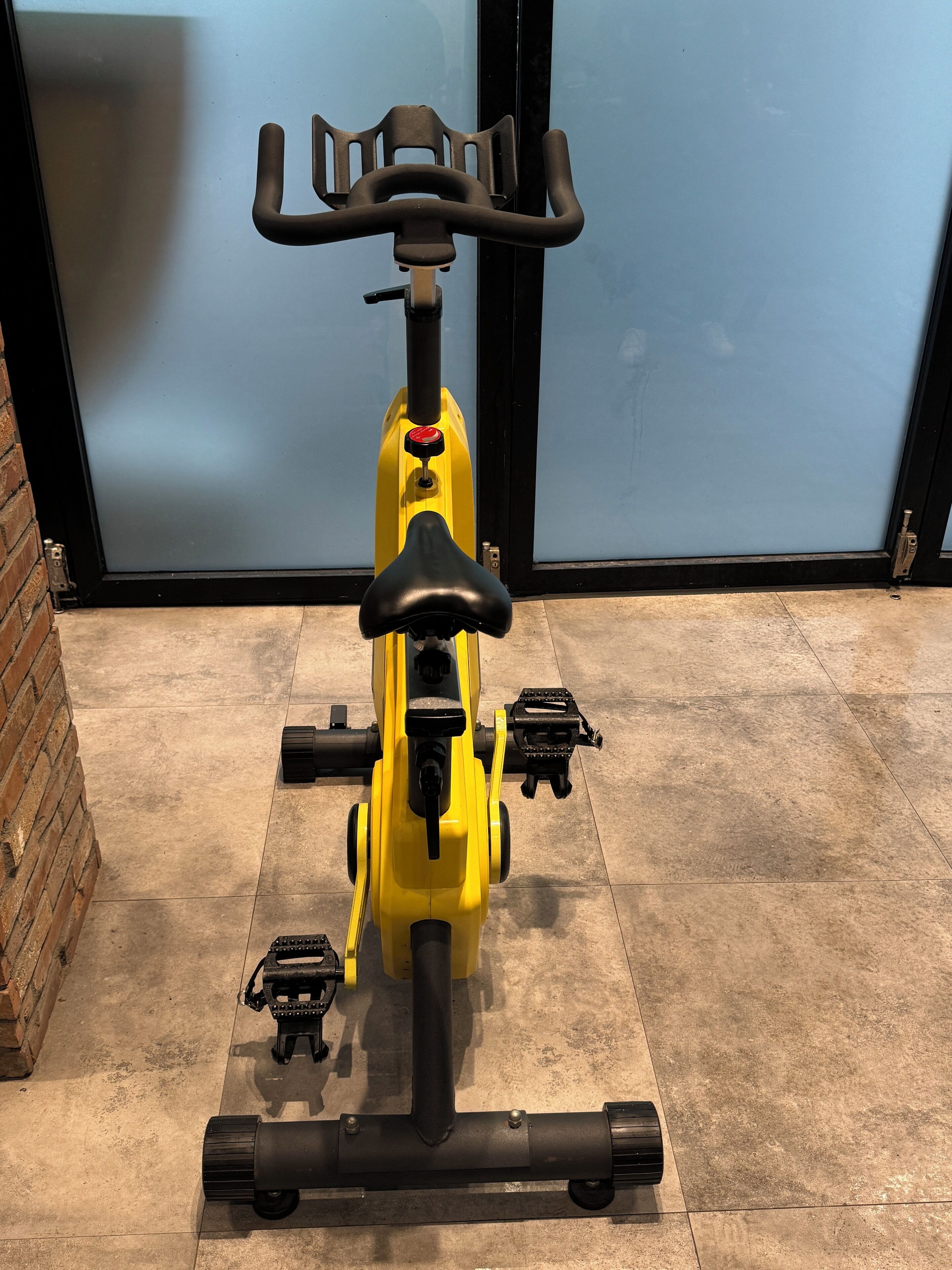 Una bicicleta estática con estructura amarilla, asiento negro y manillares, ubicada sobre un suelo de baldosas con una pared de ladrillo y una puerta de vidrio al fondo.