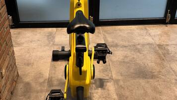 Una bicicleta estática con estructura amarilla, asiento negro y manillares, ubicada sobre un suelo de baldosas con una pared de ladrillo y una puerta de vidrio al fondo.