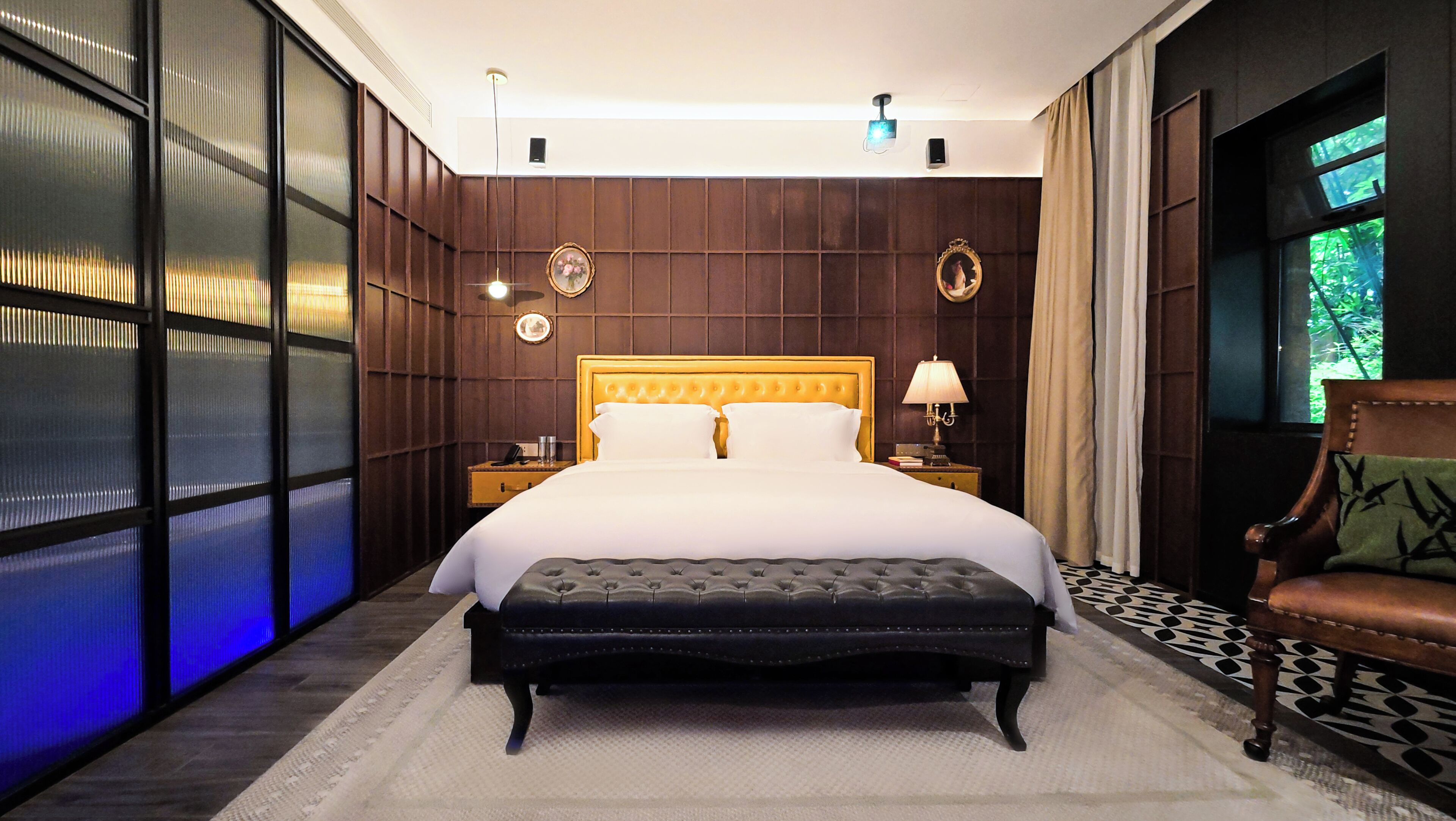 Une chambre d’hôtel avec un grand lit, une tête de lit en bois, une table de chevet avec une lampe, un banc et une vue sur la verdure par la fenêtre.