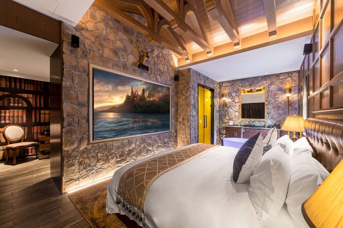 Een slaapkamer met een groot bed, stenen muren, een schilderij van een kasteel, een boekenkast en een badkamer die door een open deur zichtbaar is.