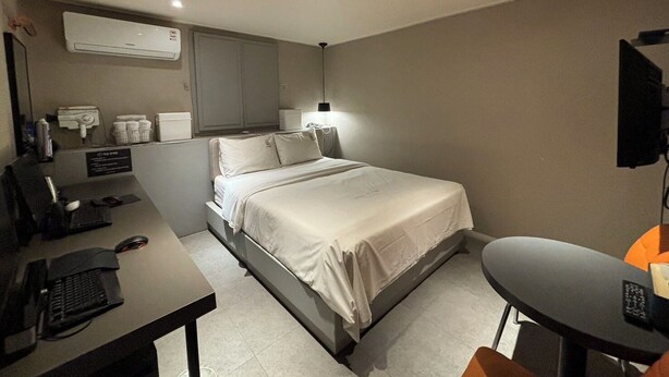 Habitación de hotel con cama, escritorio y silla. Hay una lámpara de escritorio, un tel éfono y un televisor sobre un soporte.