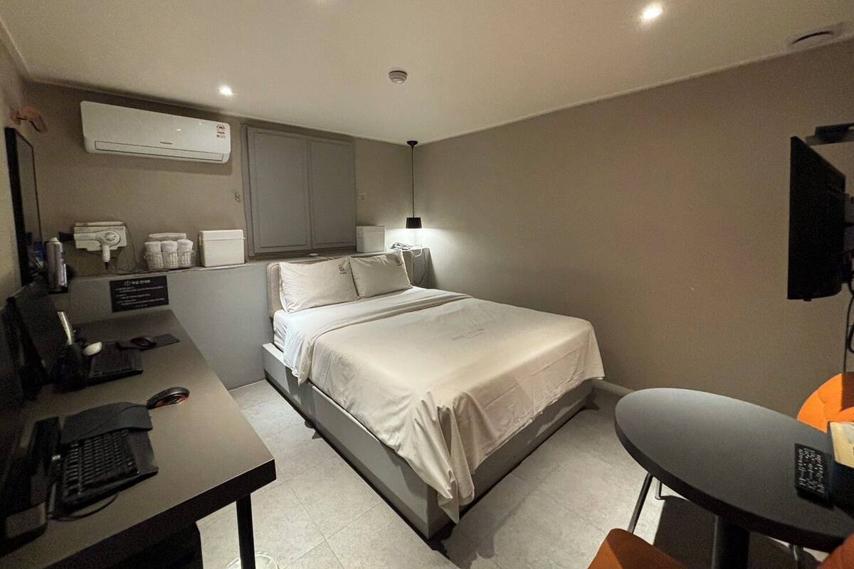 A hotel room with a bed, desk, and chair. There is a desk lamp, a phone, and a TV on a stand.