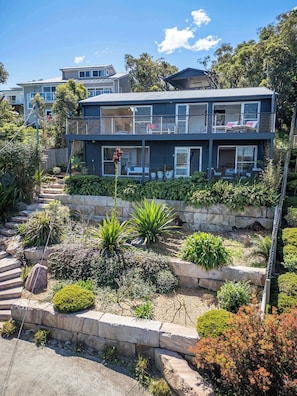 A modern two-story house with a balcony, a stone retaining wall, and a variety of plants.