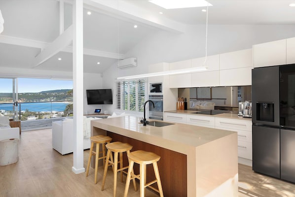 A modern kitchen with a large island, bar stools, a built-in oven, and a view of the sea.