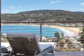 A balcony with a wooden chair and a view of a beach and hillside.