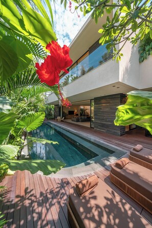 A modern outdoor pool area with a wooden deck, lounge seating, and lush greenery.