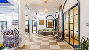 A hotel lobby with a checkered floor, a hanging spherical chair, a sofa with striped cushions, and a large window with a view.