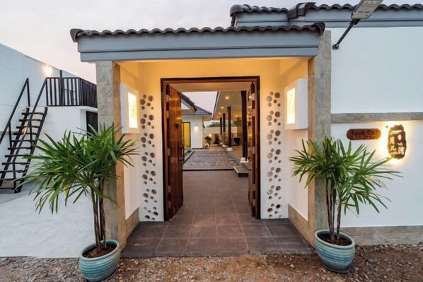 A house entrance with a tiled pathway, potted plants, a staircase, and a well-lit interior visible through the doorway.