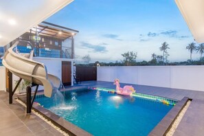 Una piscina con tobogán, un flotador de flamenco rosa y vista a palmeras.