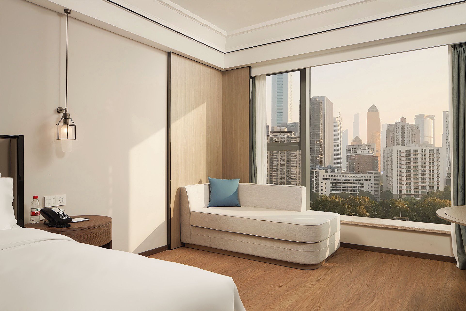 A hotel room with a bed, a seating area with a cushion, a bedside table with a phone and water bottle, a wall-mounted light fixture, and a large window with a city view.