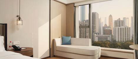 Habitación de hotel con cama, zona de estar con cojín, mesita de noche con teléfono y botella de agua, lámpara de pared y un ventanal con vista a la ciudad.