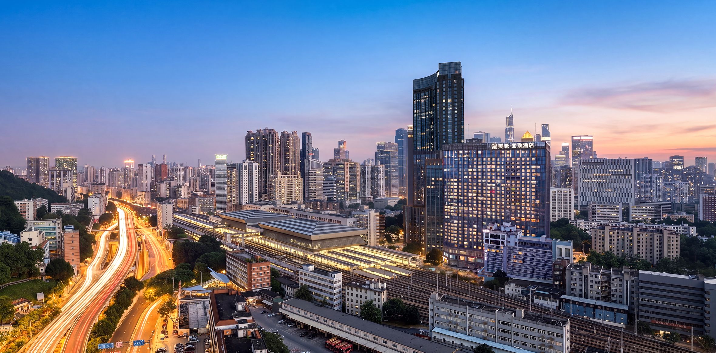 A cityscape with a prominent modern building featuring a unique curved design, surrounded by other high-rise buildings and a well-lit train station.