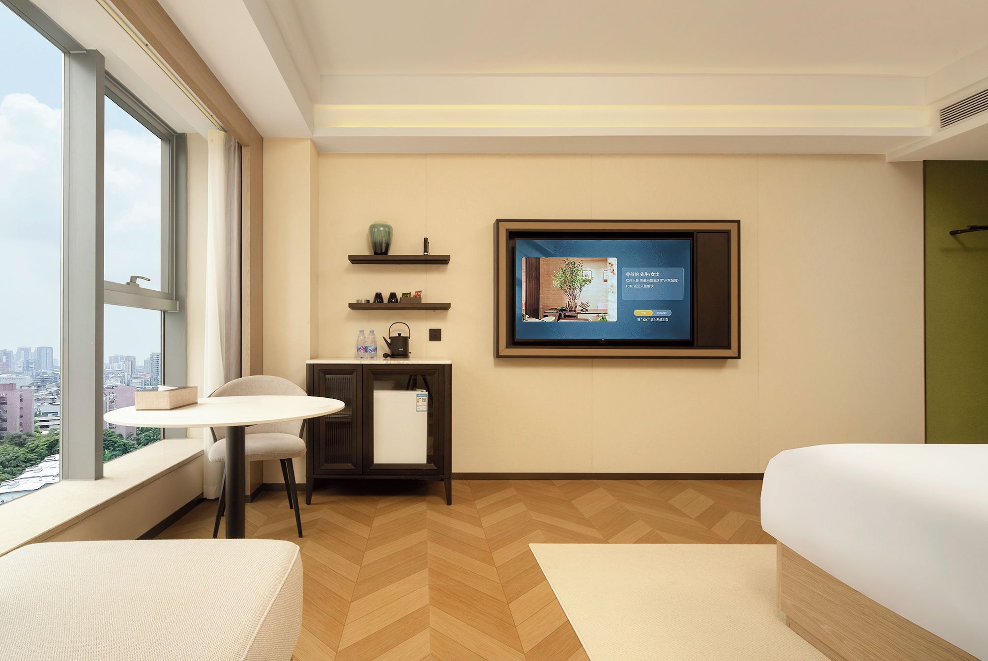 A hotel room with a large window, a TV mounted on the wall, a small table with a chair, and a bed.