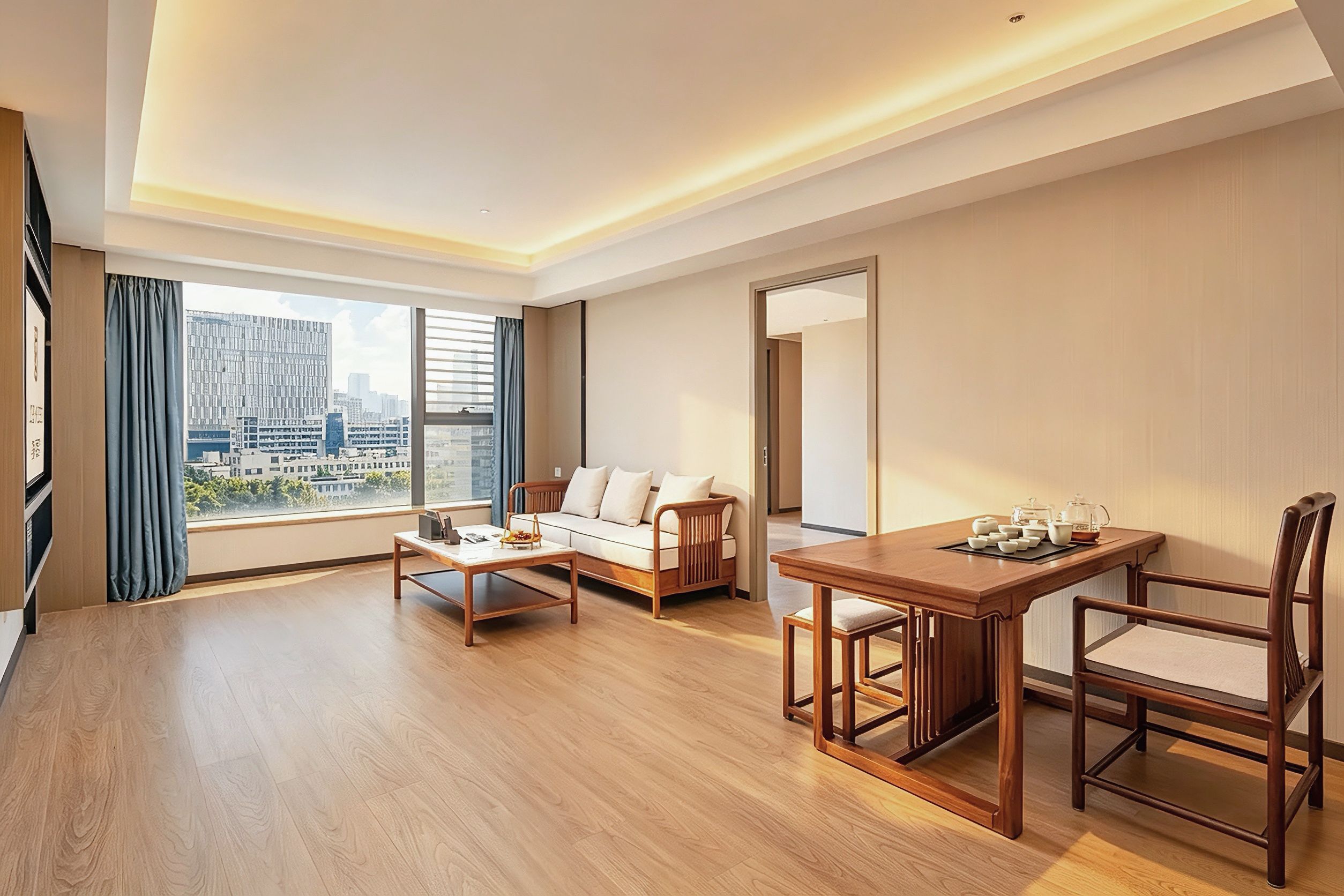 A modern hotel room with a sofa, a dining table with chairs, a large window with a city view, and a mirror on the wall.