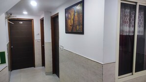 A hallway with a tiled floor, a door with a number plate, a wall-mounted painting, and a window with patterned curtains. - HOTEL AVSK (New Delhi)