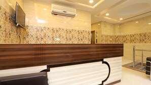 A reception area with a marble wall, a wooden reception desk, a flat-screen TV, and a ceiling-mounted air conditioner. - HOTEL AVSK (New Delhi)