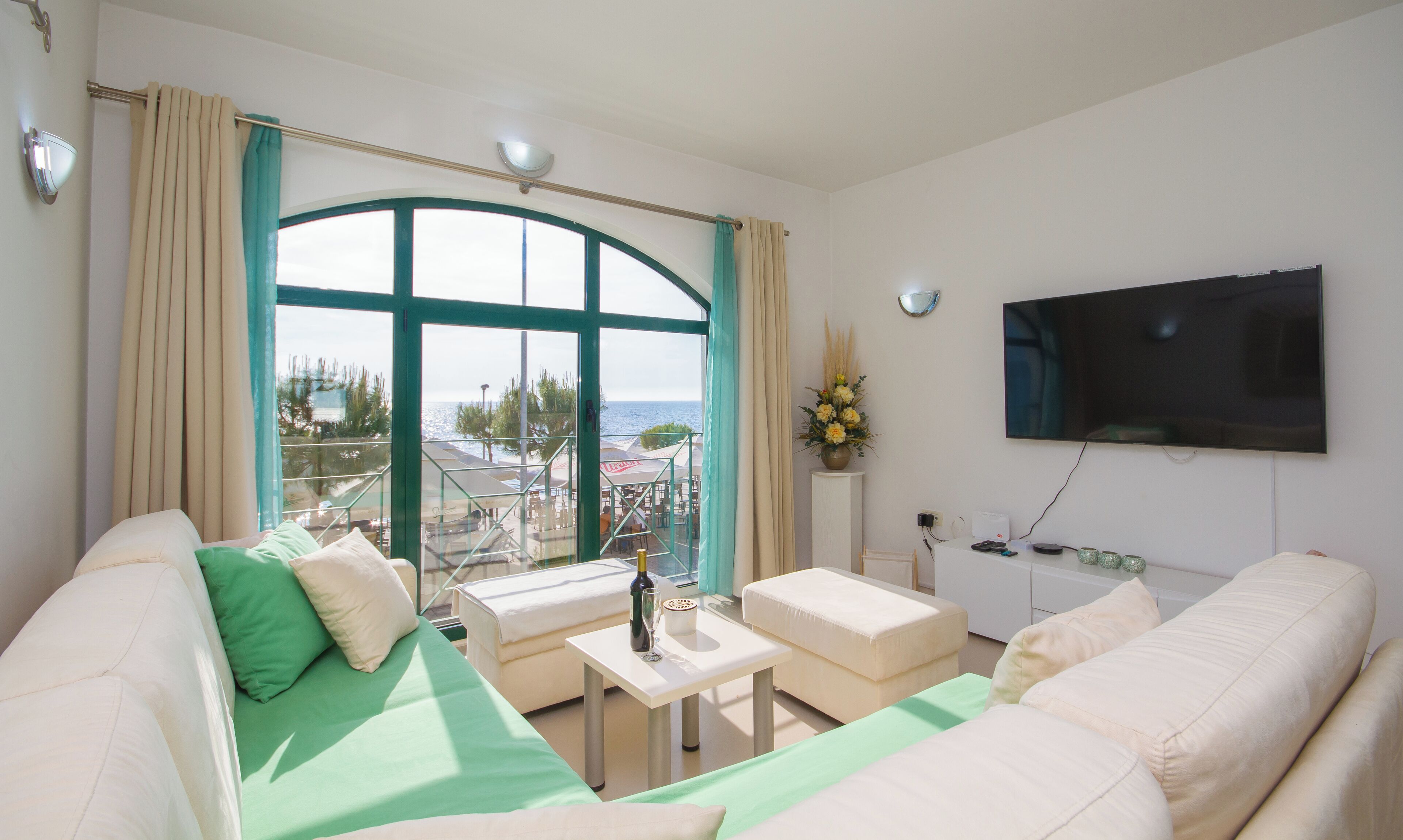 A modern living room with a large window, a flat-screen TV, and a balcony with outdoor seating.