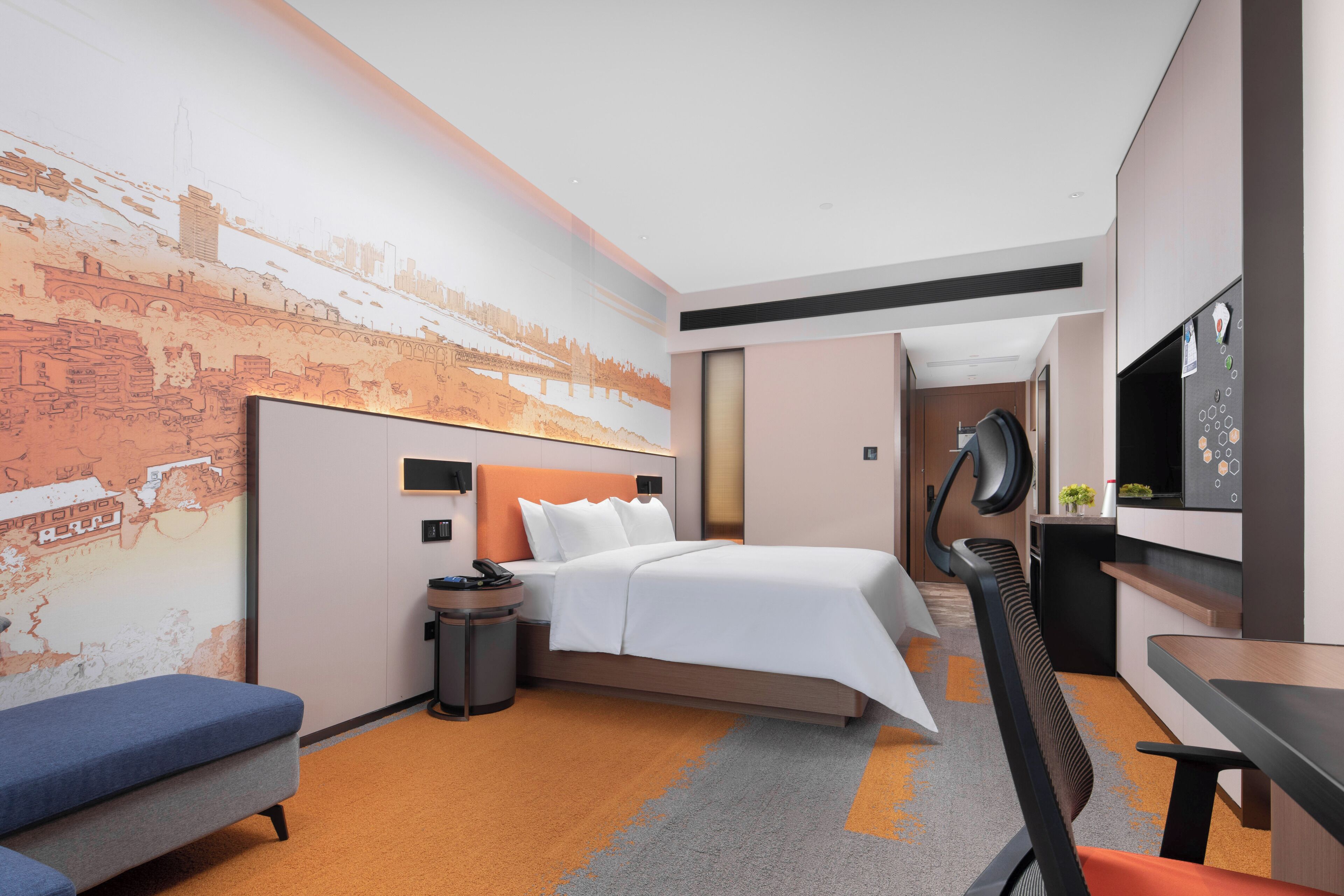A modern hotel room with a bed, a desk, a chair, a small table, a trash bin, and a wall with a cityscape mural.