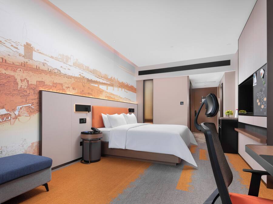 Una habitación de hotel moderna con cama, escritorio, silla, mesita, cesto de basura y una pared con un mural de un paisaje urbano.