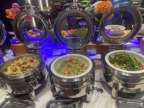 Un bufé con diversos platos en recipientes metálicos calientes, incluyendo ensaladas y salteados.