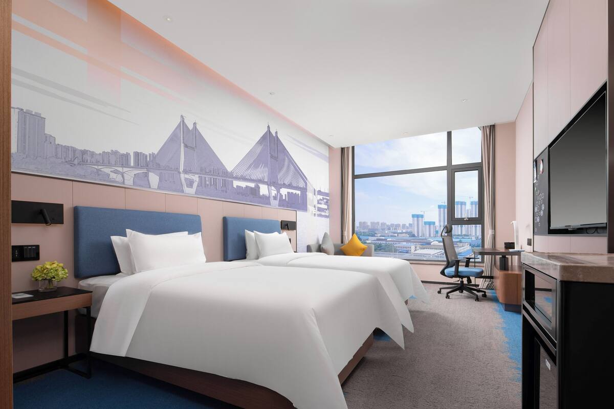 Habitación de hotel con dos camas, un escritorio, una silla, un televisor y un ventanal con vistas a la ciudad.