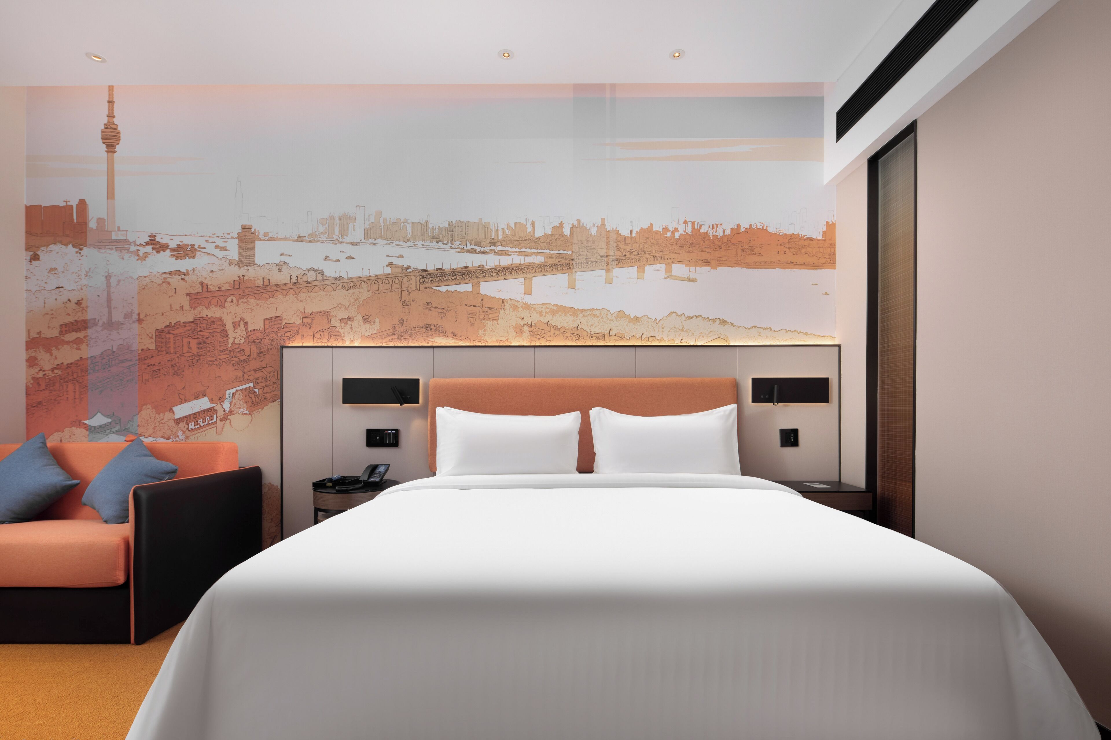 A modern hotel room with a large bed, a sofa, and a cityscape mural on the wall.