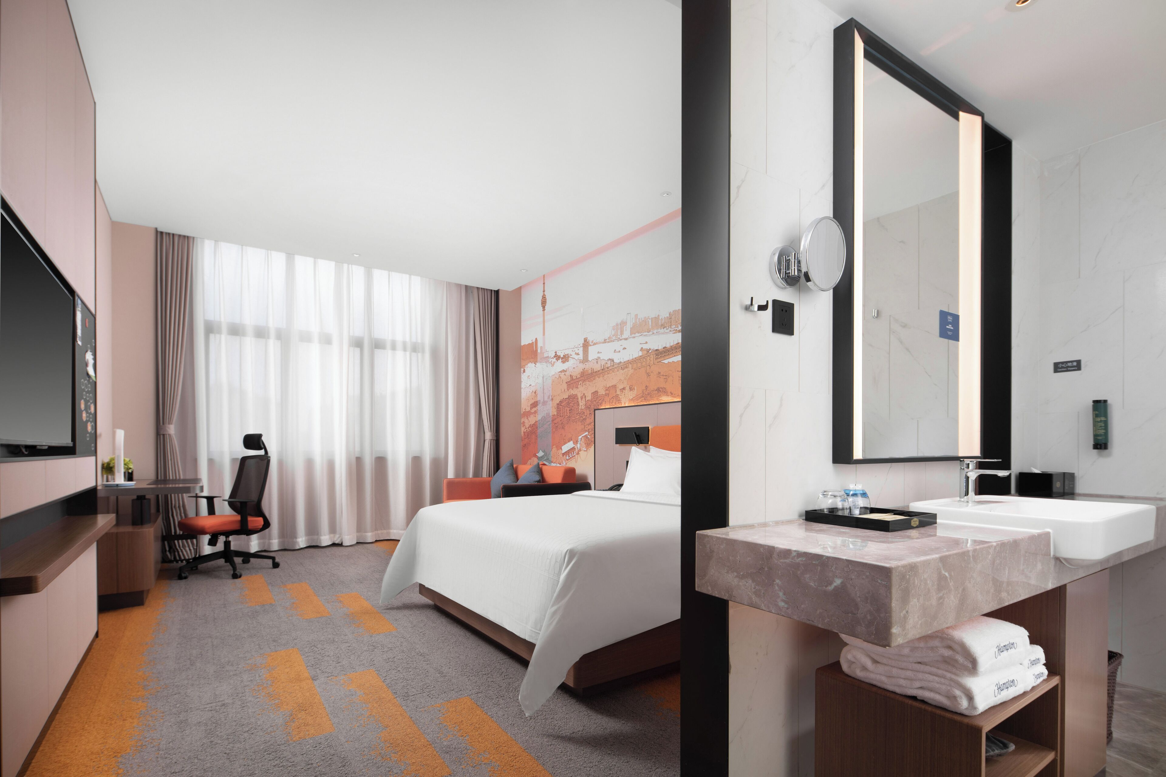 A hotel room with a bed, a desk, a chair, a large mirror, and a bathroom with a sink and towel rack.