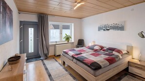 Une chambre avec un plafond en bois, un lit recouvert d’une courtepointe, une table de chevet, une chaise, une fenêtre donnant sur un beau paysage et un radiateur.