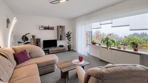 Un salon moderne avec un canapé, une table basse, un téléviseur et une grande fenêtre agrémentée de plantes.