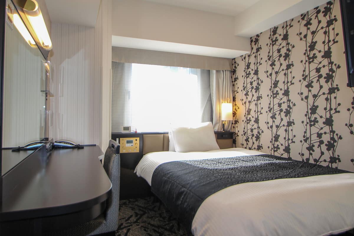 A hotel room with a bed, a desk, a chair, a nightstand, a lamp, a window with curtains, and a wall with a floral pattern.
