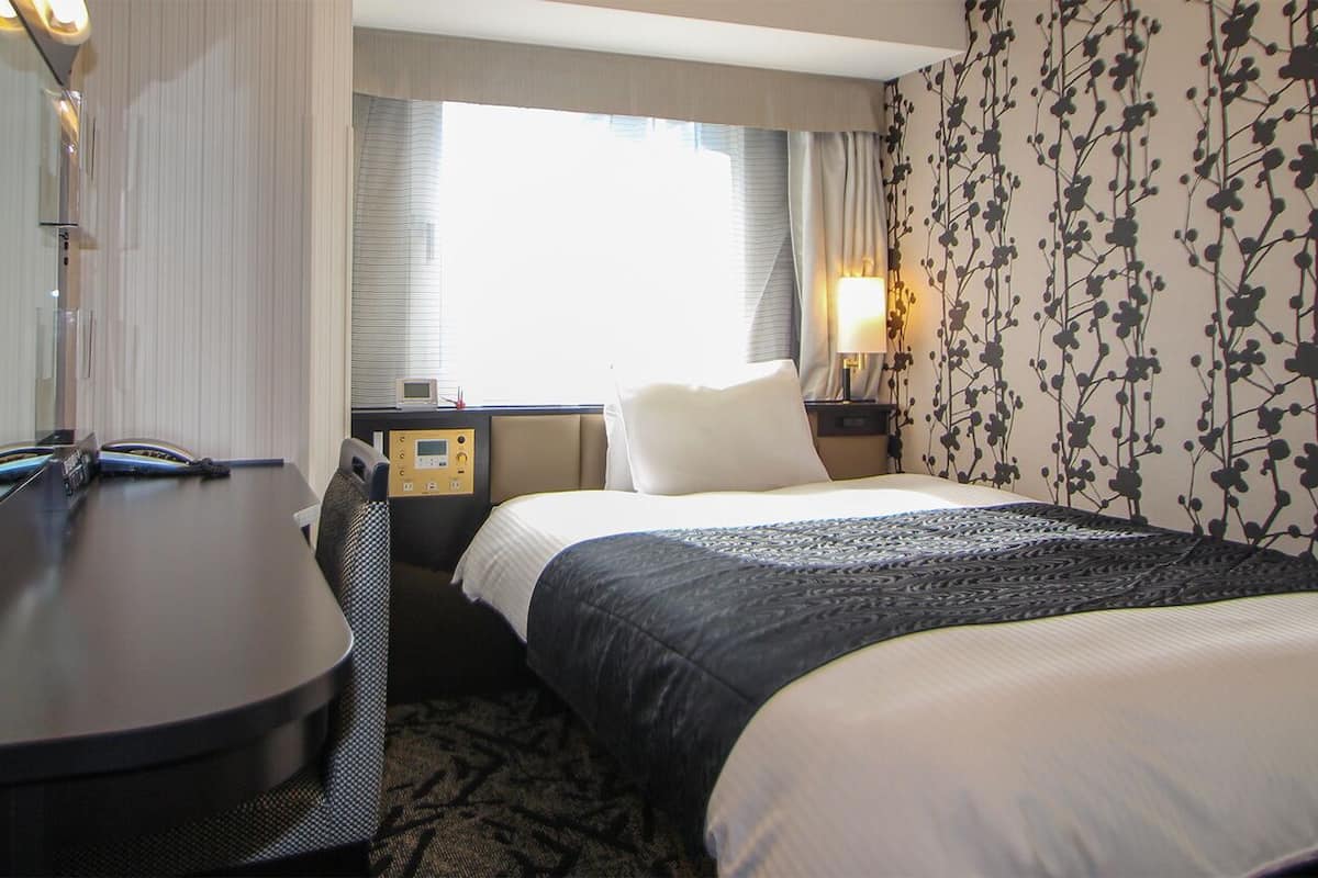 A hotel room with a bed, bedside table, lamp, and a window with curtains.